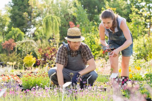 A gardener and a young girl are working together in a vibrant, well-maintained outdoor garden with a variety of flowering plants and lush green trees in the background. The gardener, wearing a straw hat, plaid shirt, and gardening apron, is kneeling on the grass, gently tending to a small flowering plant in a flower bed that features a diverse mix of colorful blooms. The girl, dressed in a denim dress and holding pruning shears, is leaning forward to assist, actively engaged in garden maintenance. The foreground showcases a lush, green lawn with closely clipped grass, while the flower beds are bordered by a mix of soil and mulch, creating a rich contrast with the foliage. Tall trees, including a weeping willow, provide partial shade, and the overall scene is bathed in bright, natural sunlight, indicating a clear, pleasant day. This image reflects typical garden care activities suitable for residential outdoor spaces in Hornsey, and subtly aligns with gardening services offered in the area by Landscapers Hornsey. The environment is peaceful and lively, highlighting a focus on outdoor aesthetics and natural growth in a suburban setting.