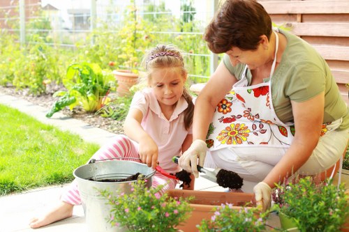 A woman and a young girl are gardening together in a well-maintained backyard, with vibrant green plants and flower beds along the fencing. The woman, wearing a floral apron and gardening gloves, is assisting the girl who is sitting on the paved patio, both engaged in planting or tending to potted flowers and shrubs. The area features a neat lawn, bordered by flower beds with a variety of plants, and a wooden fence or wall nearby. In the background, green foliage and possibly garden equipment or containers are visible. The scene is lit by natural daylight, suggesting a clear, mild-weather day. This outdoor space reflects a thoughtfully landscaped garden, suitable for family gardening activities, supported by local gardening services such as those offered by Landscapers Hornsey, focusing on lawn care, planting, and outdoor maintenance in the Hornsey area of North London.