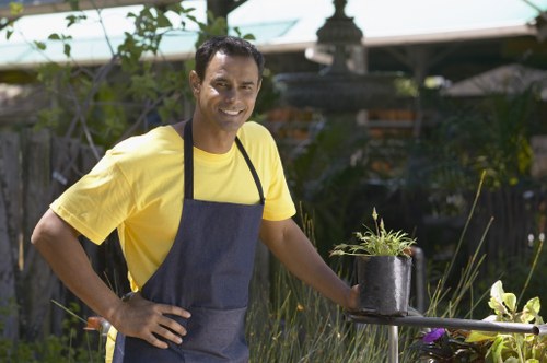 A smiling man wearing a yellow T-shirt and navy apron stands in a well-maintained garden during daytime, holding a small potted plant with green foliage. The garden features a lush grass lawn in the foreground, bordered by flower beds with blooming plants and greenery, and a wooden fence in the background. Surrounding the man are various garden elements, including shrubs and small trees, with sunlight filtering through the foliage creating a pleasant outdoor atmosphere. The scene reflects a professional gardening environment, suitable for landscaping and lawn care services, aligning with the offerings of landscapers in Hornsey and the surrounding area. The garden has a tidy, landscaped appearance, indicating regular maintenance and care, supporting the services detailed on the Hornsey patio repair solutions page.
