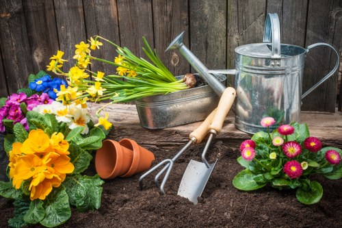 A neatly maintained garden bed in a residential outdoor space featuring dark, freshly turned soil ready for planting. The garden is bordered by a weathered wooden fence, providing privacy and a rustic backdrop. Prominent in the scene are vibrant flowering plants, including pink and yellow primroses with lush green leaves, and a cluster of yellow and blue daffodils. Gardening tools such as a small hand rake, trowel, and a hand fork are resting in the soil, indicating recent planting activity. To the right, there is a silver watering can and a terracotta flowerpot lying on its side, suggesting ongoing garden care. Additionally, there are three small, empty terracotta pots stacked near the flowers. The overall setting conveys an outdoor garden area in preparation for new planting, suitable for services like lawn care and garden maintenance by companies such as Landscapers Hornsey, in the Hornsey area, N8 post code. Natural daylight light illuminates the scene, emphasizing the vibrant plant colours and the textures of the soil and garden tools.