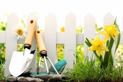 A close-up view of a gardening scene set in a front garden or backyard with a white picket fence in the background, featuring a patch of lush, green grass and vibrant yellow daffodil flowers blooming on the right side. In the foreground, there are gardening tools including a small trowel with a wooden handle, a cultivator with a teal handle, and a pair of gardening gloves placed on the ground, suggesting recent or ongoing garden maintenance. The scene is illuminated by natural daylight, and the background foliage is softly blurred, emphasizing the garden elements and tools. The setting reflects typical outdoor gardening activities that a professional landscaper like Landscapers Hornsey might undertake as part of garden renovation and landscaping services in the Hornsey area, with the overall environment appearing well-maintained and inviting for outdoor improvement.