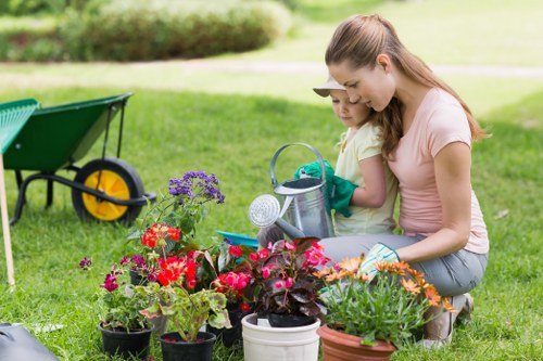 In a well-maintained outdoor garden space, a woman and a young girl are engaged in planting activities among a diverse array of flowering plants and greenery. The foreground features several potted flowers with vibrant red, pink, purple, and orange blossoms, arranged on the lush green grass. Behind them, there is a partial view of garden tools, including a green wheelbarrow, which indicates ongoing gardening work. The background displays a neatly trimmed hedge and a gently sloping lawn that extends into a softly blurred, natural landscape, suggesting a peaceful residential garden environment typical for outdoor lawn care and landscaping services in Hornsey. The scene is illuminated by bright, natural daylight, highlighting the fresh, natural tones of the soil, plants, and grass, reflecting a cared-for outdoor space suitable for family gardening activities. This setting demonstrates the benefits of professional gardening care from Landscapers Hornsey, helping to maintain vibrant, healthy lawns and attractive flower beds, characteristic of quality garden services in the local area.