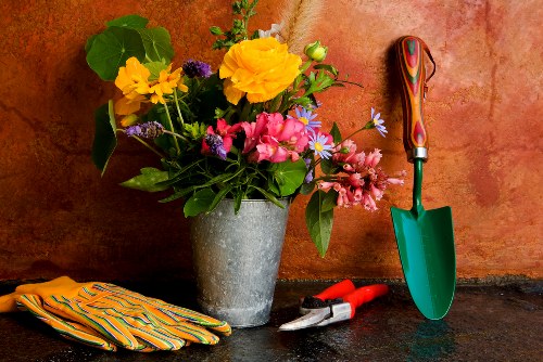 A close-up of a gardening workspace in a back garden, featuring a large, grey ceramic pot filled with a vibrant assortment of freshly cut flowers including yellow, pink, purple, and blue blooms, surrounded by green foliage. To the right of the pot, there is a small green-handled trowel with a wooden grip standing upright against the textured terracotta-colored wall, accompanied by red-handled pruning shears and a silver garden hand fork resting on the dark stone surface. On the left side, a pair of striped gardening gloves made of thick, durable material are neatly folded. The outdoor environment suggests a well-maintained garden area, with the flowers and tools indicating ongoing planting, maintenance, or floral arrangements, consistent with professional gardening and landscaping practices in the Hornsey area. The scene is illuminated by natural light, highlighting the vivid colors and textures of the floral display and garden tools, reflecting a professional approach to garden care by companies such as Landscapers Hornsey.