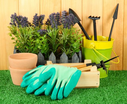 The image displays a well-organized garden storage area with various gardening tools and supplies arranged on artificial grass in a backyard or outdoor space, possibly within the Hornsey area. In the foreground, a pair of teal and green gardening gloves rests on a wooden planter box. To the left, there are multiple small terracotta plant pots stacked together, suitable for planting flowers or herbs. Behind the gloves, a rectangular plant container houses a row of lavender plants with vibrant purple flower spikes and lush green foliage, indicating a love of colourful, fragrant plants typical of UK gardens. To the right, a bright yellow plastic bucket holds gardening tools such as a hand trowel, pruning shears, and a cultivator, all positioned upright for easy access. The background features a natural wooden panel fence, suggesting a private garden setting. The scene captures a tidy, functional outdoor workspace, reflecting typical gardening activities like planting and maintenance, and showcasing elements that landscapers Hornsey might incorporate in garden landscaping projects, with natural lighting indicating a clear day.