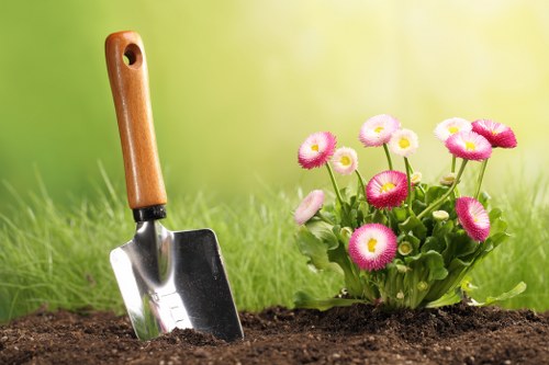 A garden scene featuring a small section of soil in the foreground, with a wooden-handled gardening trowel planted into the earth on the left side. Behind the soil, there is a vibrant, healthy flower bed filled with pink and white daisies, surrounded by lush green foliage. The background displays a blurred, naturally lit outdoor space with a grassy lawn that extends into the distance, indicating a well-maintained garden area typical of residential landscaping in Hornsey. The overall setting suggests a peaceful, cultivated outdoor environment suitable for gardening and lawn care services provided by Landscapers Hornsey, with natural tones of green, pink, and brown highlighted under soft daylight, hinting at mild weather conditions common in early autumn or late spring. The scene emphasizes garden planting, flower bed maintenance, and general outdoor landscape presentation consistent with professional gardening services.